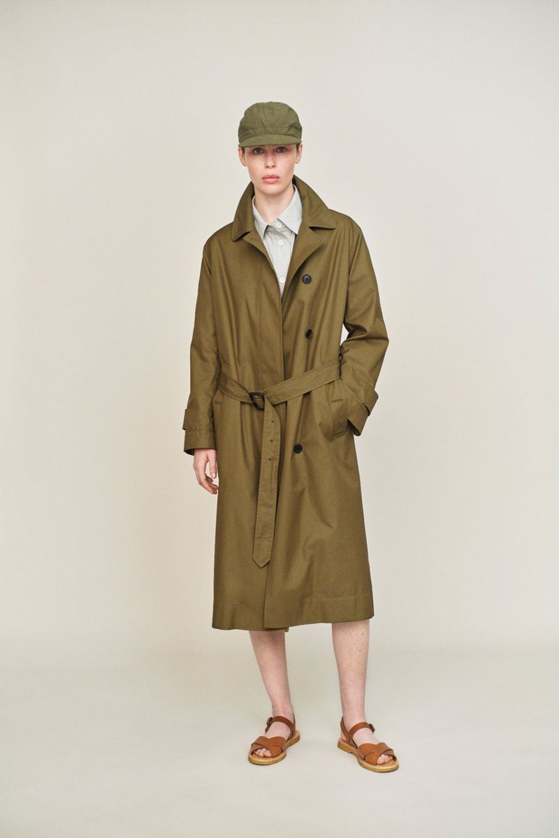 A person stands in a light studio wearing an olive trench coat, cap, light shirt, and sandals against an off-white backdrop.