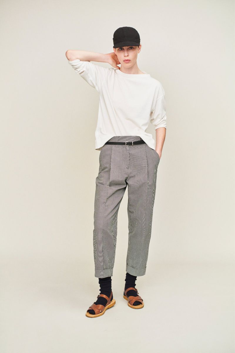 A full-length studio shot of a model posing in a white long-sleeve shirt, checkered pants, black socks, sandals, and black cap against a light background.