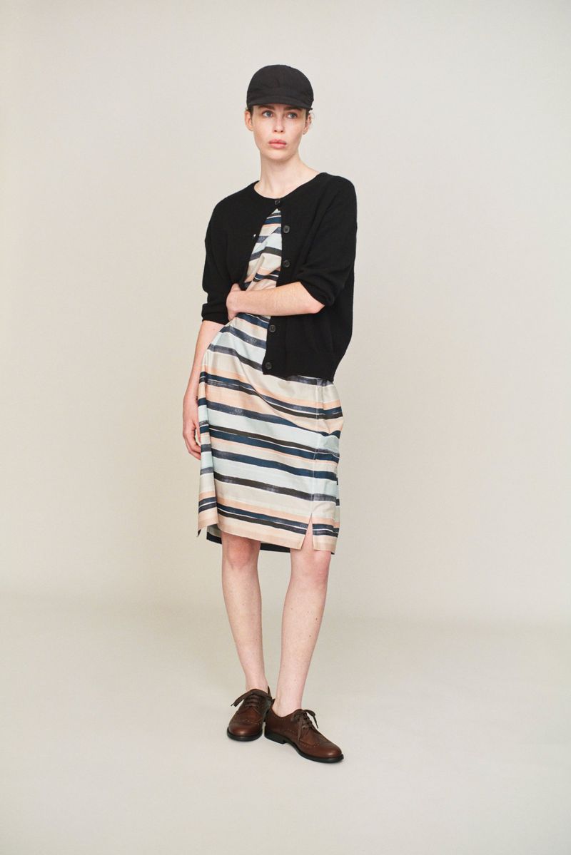 Studio shot of an adult woman wearing a striped dress, black cardigan, cap, and dark brown shoes, standing against a neutral backdrop.
