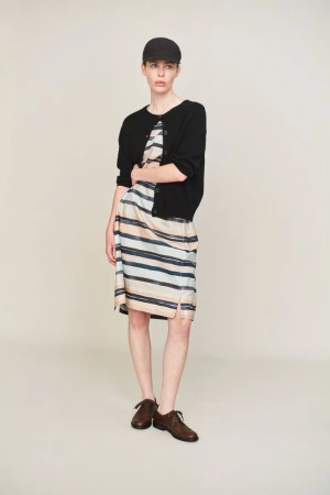 Studio shot of an adult woman wearing a striped dress, black cardigan, cap, and dark brown shoes, standing against a neutral backdrop.