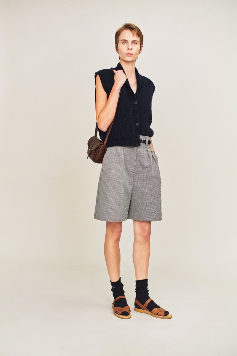 Full shot of a slender person in vest, shorts, socks, and sandals with bag, standing against a neutral background in a fashion style.