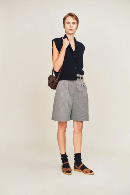 Full shot of a slender person in vest, shorts, socks, and sandals with bag, standing against a neutral background in a fashion style.