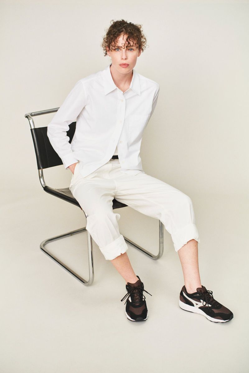 A young adult woman with curly hair sits in a black chair wearing a white button-down shirt, white pants, and black athletic shoes in a studio.