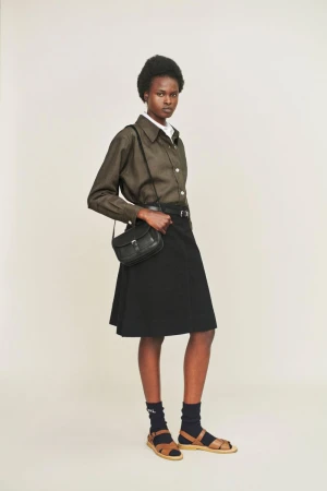 Model wearing an olive green shirt, black skirt, and leather crossbody bag standing against a white seamless backdrop.