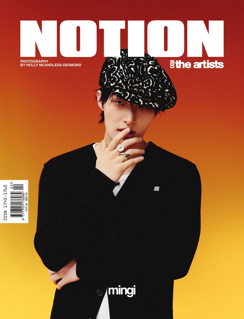 Magazine cover portrait of a man wearing a patterned flat cap and black cardigan against a warm gradient background.