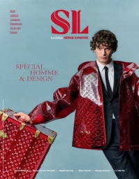 A man wearing a suit jacket and red patterned coat poses next to a matching red trunk against a blue background.