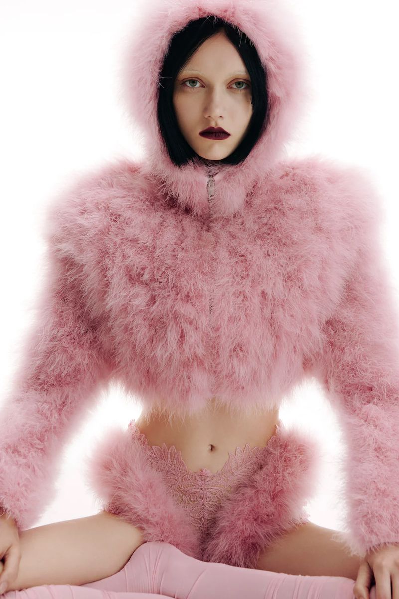 Fashion model in a light pink feathered outfit with a hood, posing in a light studio setting. Dark lipstick, sleek black hair, fair skin.