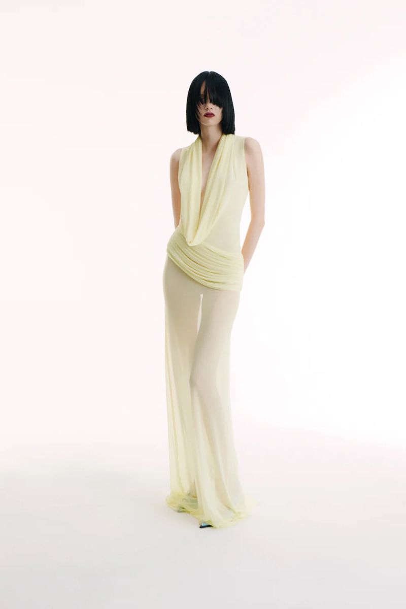A slender woman with dark hair poses in a pale yellow sleeveless top and sheer pants against a white backdrop in a fashion shot.