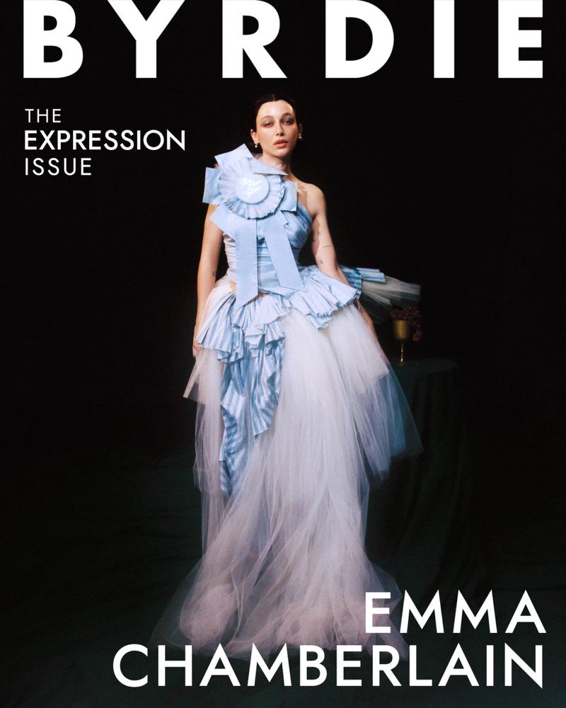 Emma Chamberlain poses on a Byrdie magazine cover in an elaborate blue gown with tulle, standing in front of a dark studio backdrop.