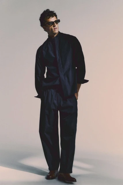 Full-length portrait of a male model in a relaxed pose, wearing a dark blue matching shirt and pants outfit with brown dress shoes.