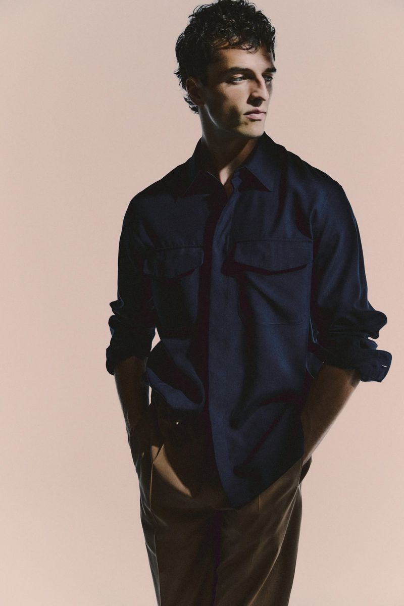 A young adult man poses in a navy blue shirt and brown pants, looking off to the side in a brightly lit photo studio setting.