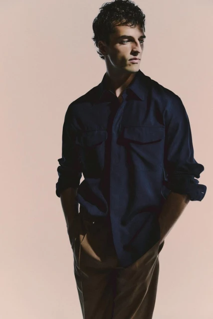 A young adult man poses in a navy blue shirt and brown pants, looking off to the side in a brightly lit photo studio setting.