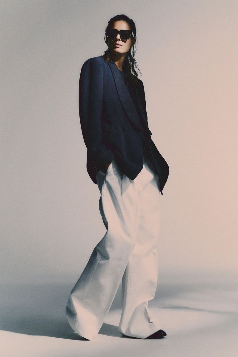 Model wearing dark sunglasses, a dark blazer, and white trousers against a neutral background