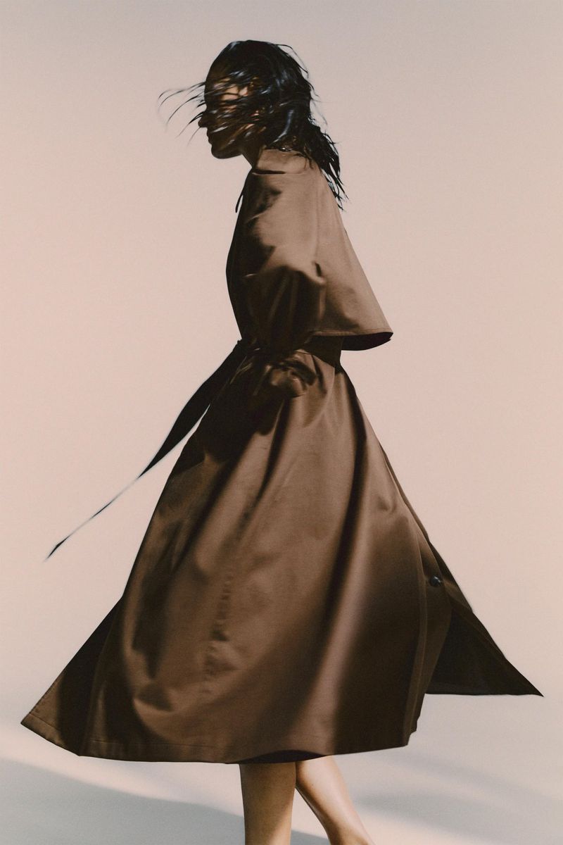 Model in motion wearing a long brown belted coat against a plain light background