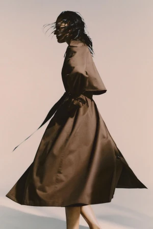 Model in motion wearing a long brown belted coat against a plain light background