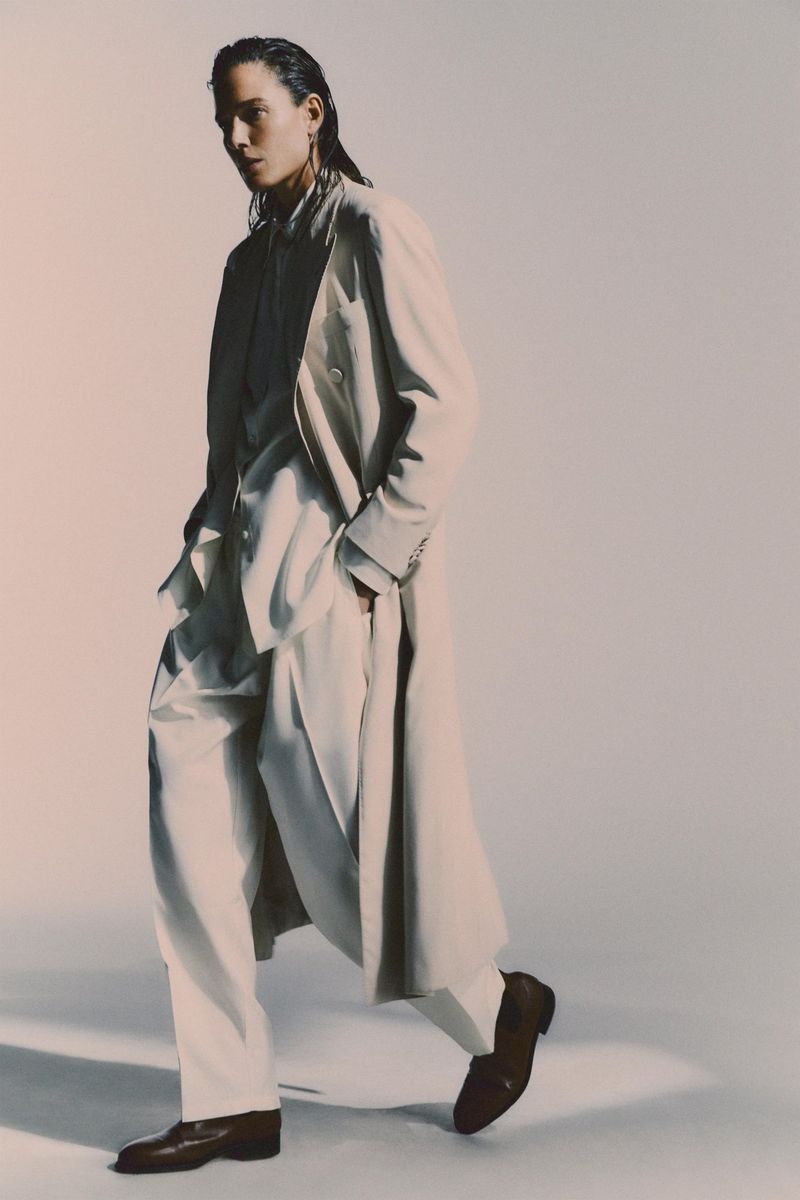 Stylish man in a light overcoat and loose pants walking against a neutral backdrop, showcasing modern fashion and confident stride.