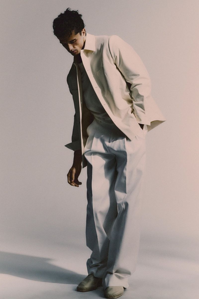 Male model in light-colored, relaxed-fit shirt and trousers, standing in a photo studio with a slight lean and hands casually in pockets.