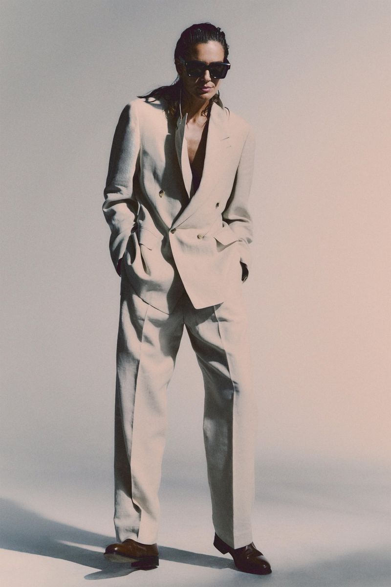 An adult male poses in a light-beige double-breasted suit with dark shoes against a light-beige gradient backdrop in a fashion shot.