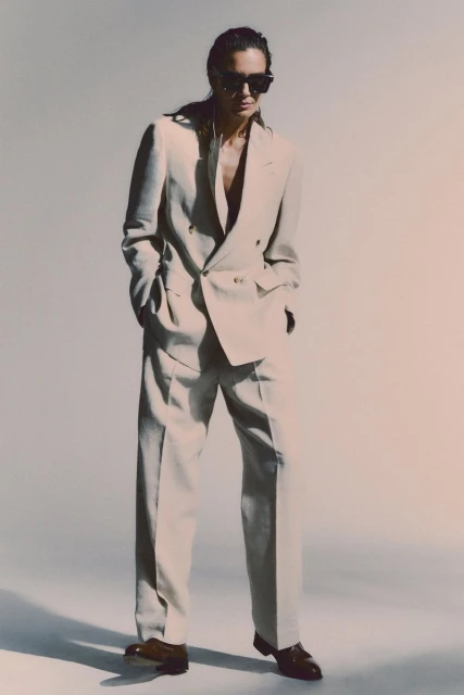 An adult male poses in a light-beige double-breasted suit with dark shoes against a light-beige gradient backdrop in a fashion shot.