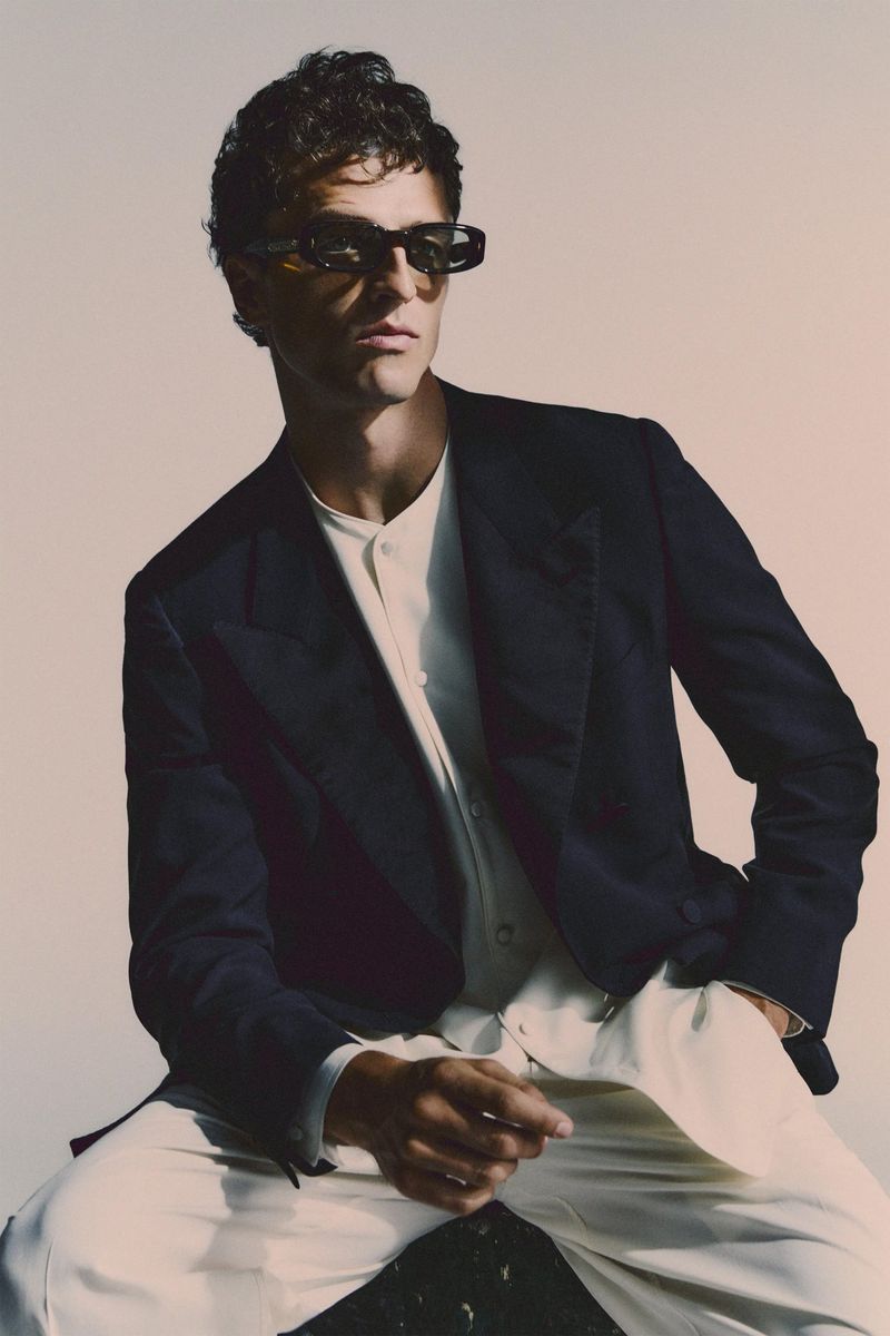 Portrait of a man wearing sunglasses and a navy blazer, posing indoors against a neutral backdrop. He has curly hair and is seated.