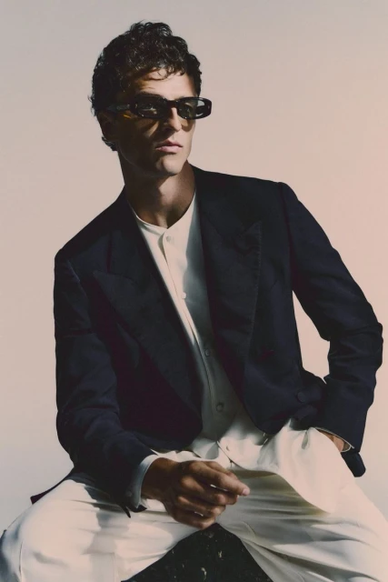 Portrait of a man wearing sunglasses and a navy blazer, posing indoors against a neutral backdrop. He has curly hair and is seated.