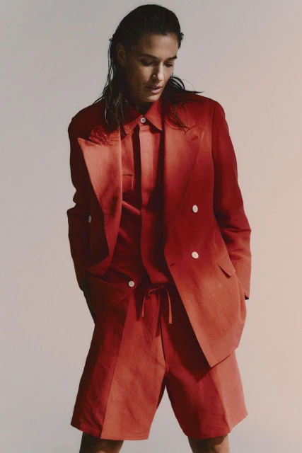 Adult woman poses in a reddish-brown blazer, button-down shirt, and shorts against a neutral background in a fashion editorial shot.