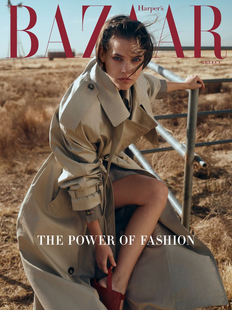 Harper's Bazaar cover with a stylish woman in a trench coat posing on a metal structure, set against a rural backdrop.