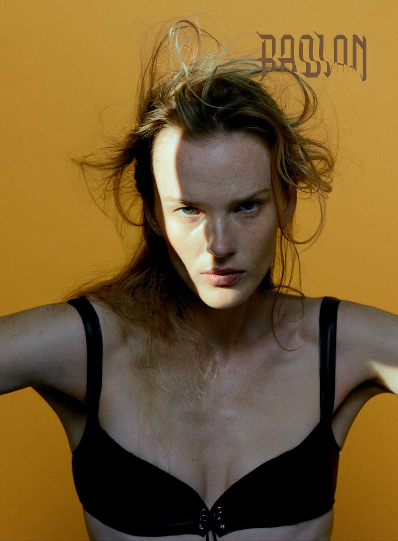 Portrait of a fair-skinned woman with long blonde hair and a black bra against a yellow backdrop, with the word 'PASSION' on top.