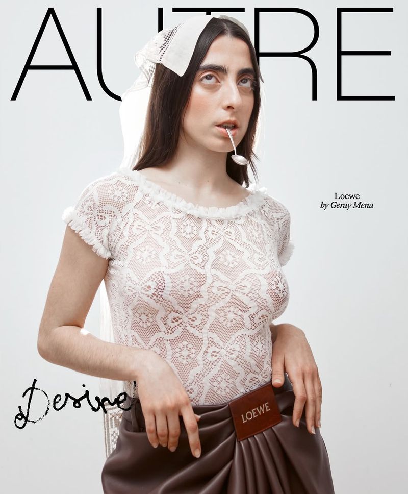 A woman with medium-length dark hair and a lace top poses in a studio with a spoon in her mouth for a fashion editorial.