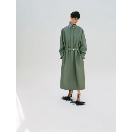 Model wearing a sage green belted coat and fringed heels against a seamless white studio background.