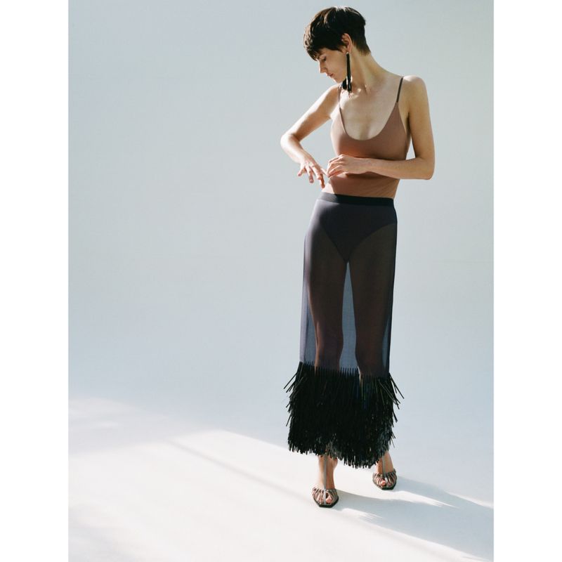 A slender woman with short brown hair poses in a light brown top and sheer black skirt with tassels against a white backdrop.