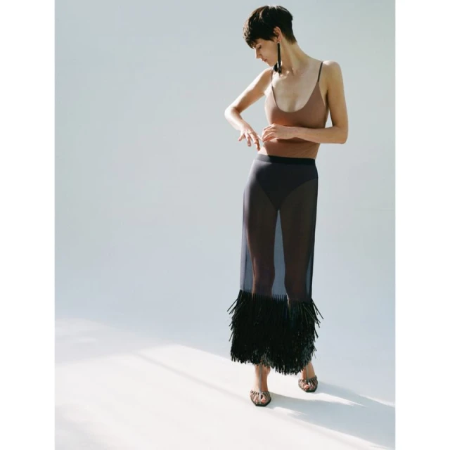 A slender woman with short brown hair poses in a light brown top and sheer black skirt with tassels against a white backdrop.