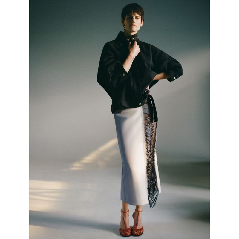 A full-length shot of a woman with short brown hair posing in a black jacket, white skirt with fringe, and high heels against a grey backdrop.