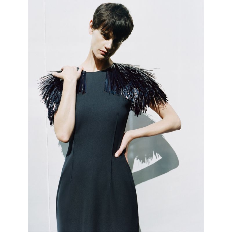A slender woman with short dark hair poses indoors wearing a black dress with a fringed shoulder embellishment against a white wall.