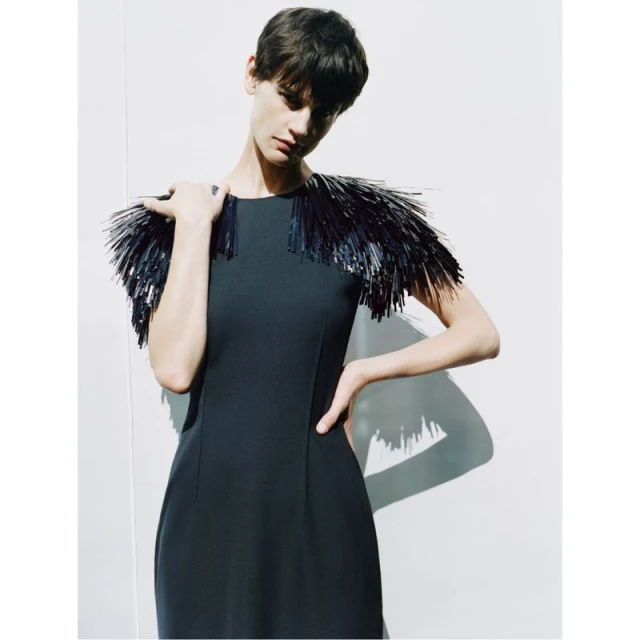 A slender woman with short dark hair poses indoors wearing a black dress with a fringed shoulder embellishment against a white wall.