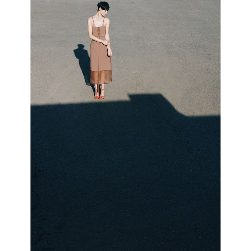 Model wearing a brown midi dress standing on an expansive asphalt surface with strong shadows
