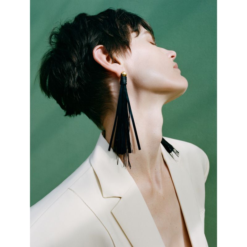 Portrait of a woman wearing a cream-colored blazer and statement black tassel earrings against a green backdrop.