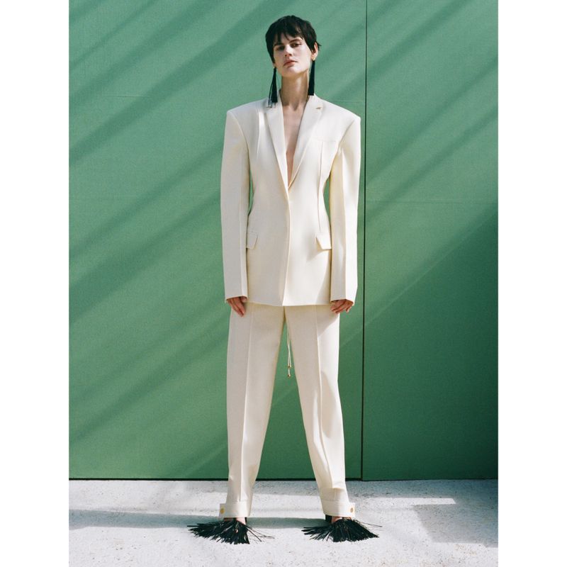Woman in a white suit standing in front of a green wall wearing black statement earrings and black tassel shoes in studio lighting.