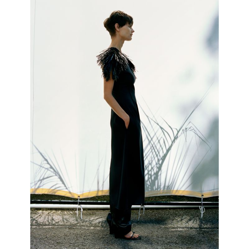 Elegant woman in a black jumpsuit with feather details stands in profile against a neutral backdrop with subtle plant shadows.