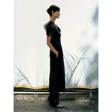 Elegant woman in a black jumpsuit with feather details stands in profile against a neutral backdrop with subtle plant shadows.