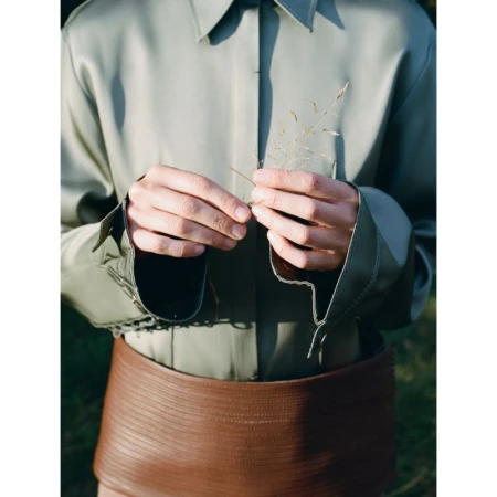 Close-up portrait of a model wearing a sage green silk shirt and a brown leather skirt holding a dry plant sprig in hands