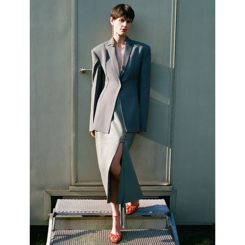 Fashion editorial shot of a woman in a grey blazer and skirt with a slit, standing on metal steps in front of a grey wall.