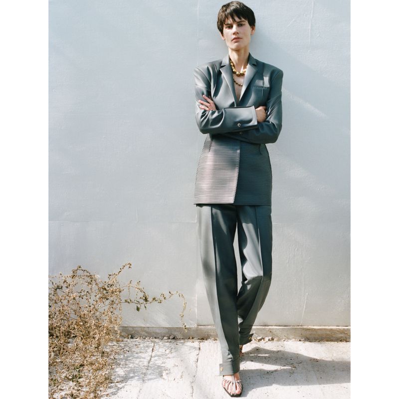Elegant woman in a grey suit poses against a white wall with vine detail, looking confidently ahead in a fashion editorial shot.