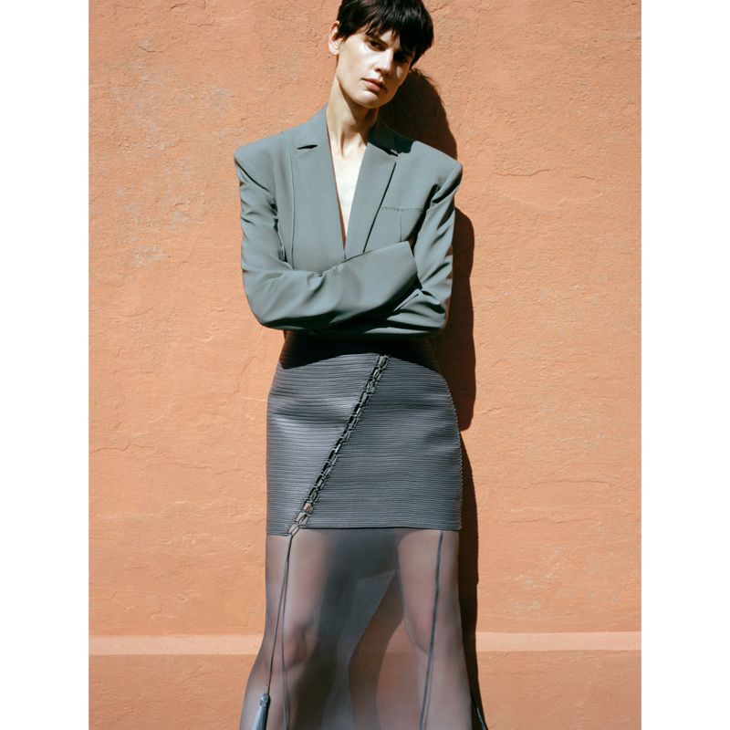 A fashionable adult woman in a blazer and skirt stands outdoors with arms crossed, posing against an orange stucco wall in soft daylight.