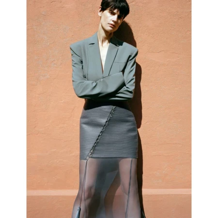A fashionable adult woman in a blazer and skirt stands outdoors with arms crossed, posing against an orange stucco wall in soft daylight.