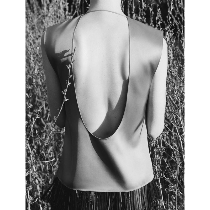 A woman with bare back poses outdoors wearing a minimalist top; the shot is in black and white and is set against a natural, leafy backdrop.