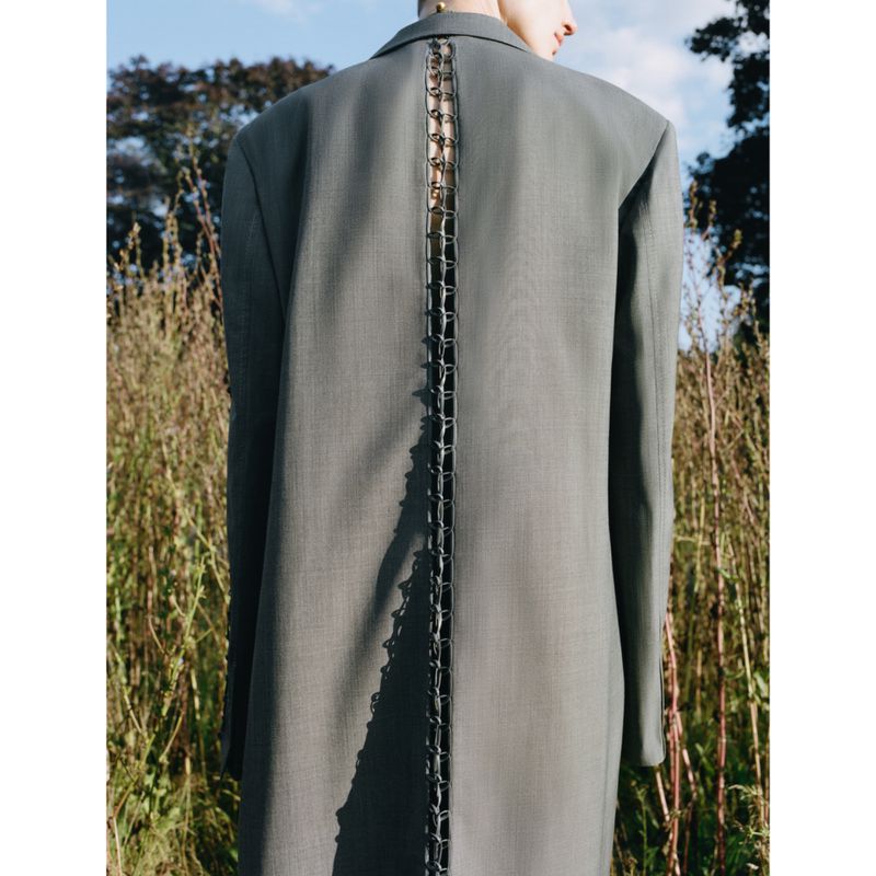Back view of an adult woman in a gray blazer, standing in a field of tall grass with a blue sky overhead in an outdoor fashion shot.