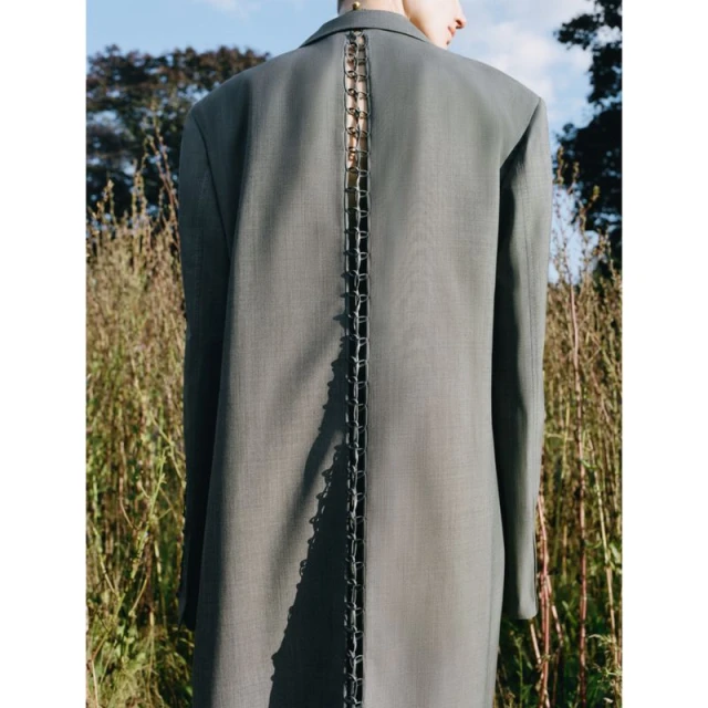 Back view of an adult woman in a gray blazer, standing in a field of tall grass with a blue sky overhead in an outdoor fashion shot.