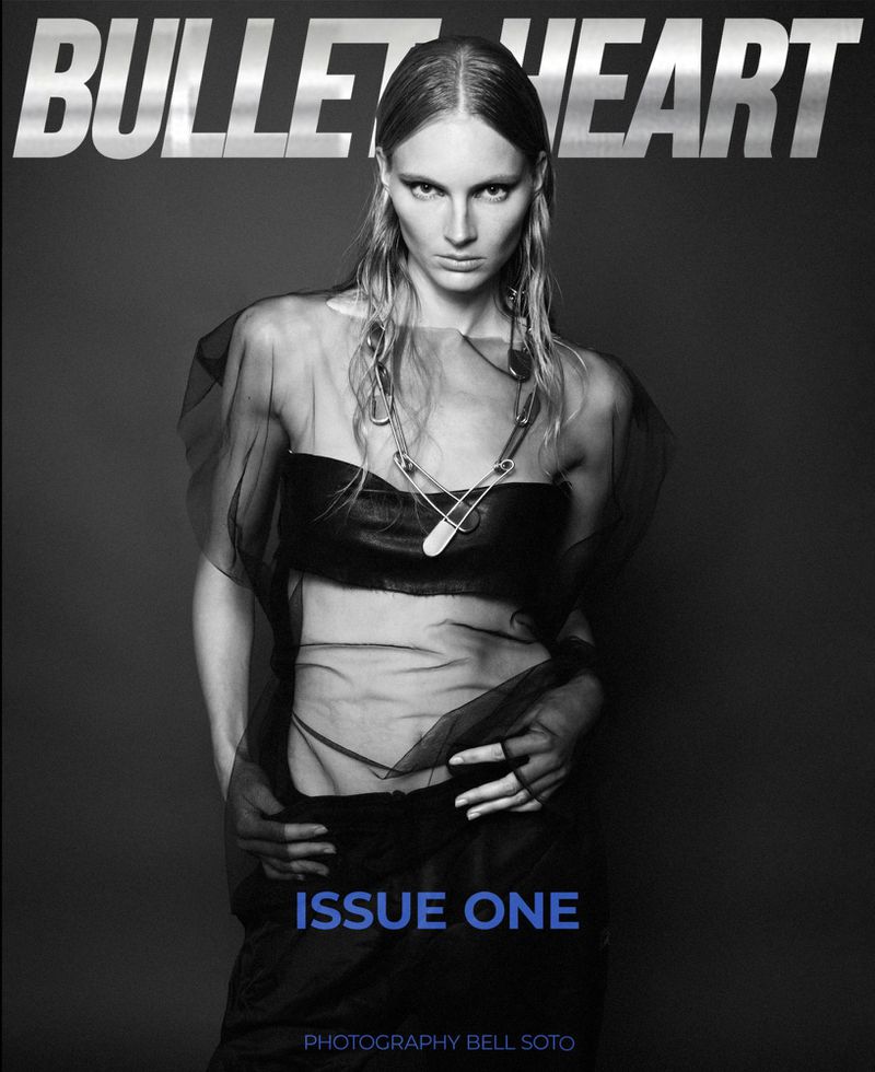 Black-and-white fashion magazine cover featuring a female model wearing a dark sheer top and large safety pin necklace, against a gray background.