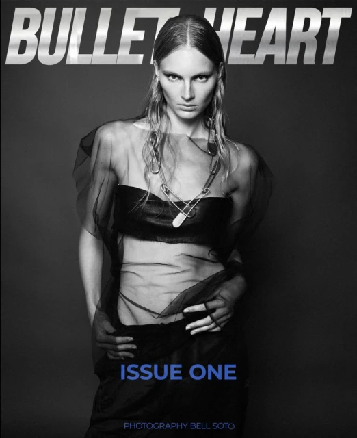 Black-and-white fashion magazine cover featuring a female model wearing a dark sheer top and large safety pin necklace, against a gray background.